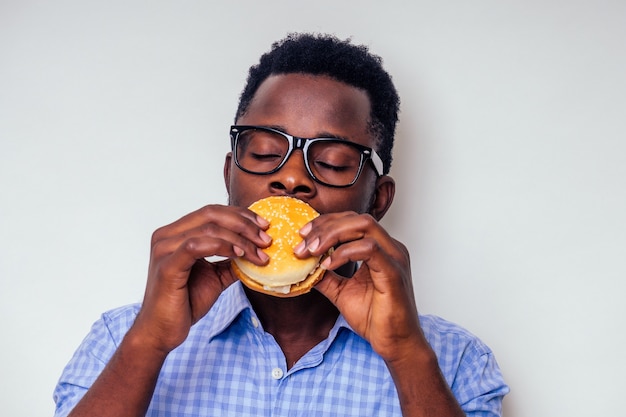 Homme afro-américain appréciant le goût du hamburger. Beau et jeune homme afro dans une chemise élégante et des lunettes tenant un hamburger sur un fond blanc. L'amour du régime de malbouffe
