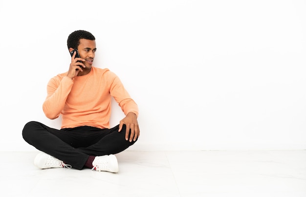 Homme afro-américain assis sur le sol sur fond de fond isolé gardant une conversation avec le téléphone portable avec quelqu'un