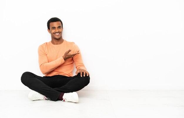 Homme afro-américain assis sur le sol sur fond de fond isolé présentant une idée tout en regardant en souriant vers