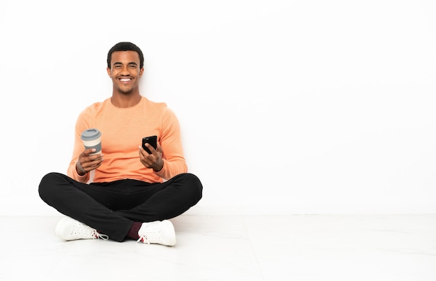 Homme afro-américain assis sur le sol sur fond de fond isolé tenant du café à emporter et un mobile