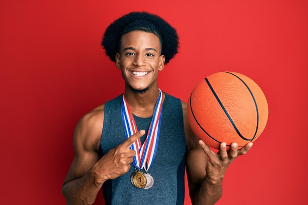Homme afro-américain aux cheveux afro portant des médailles gagnantes au basketteur souriant heureux pointant avec la main et le doigt