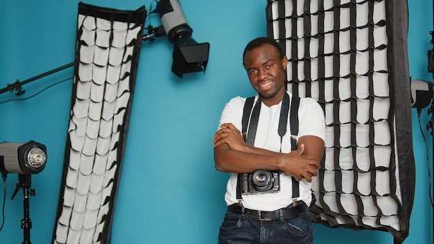 Homme afro-américain ayant un équipement de photographie dans les coulisses, utilisant un projecteur, une lampe de poche, un objectif d'appareil photo reflex numérique et un trépied. Photographe posant avec des outils créatifs en studio dans les coulisses.