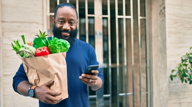 Homme afro-américain avec barbe tenant un sac en papier d'épicerie du supermarché à l'aide d'un smartphone