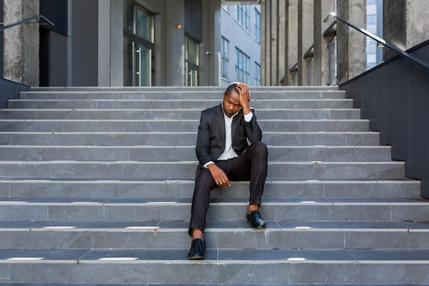 Homme afro-américain bouleversé assis dans les escaliers à l'extérieur de l'immeuble de bureaux homme d'affaires en faillite dans le désespoir