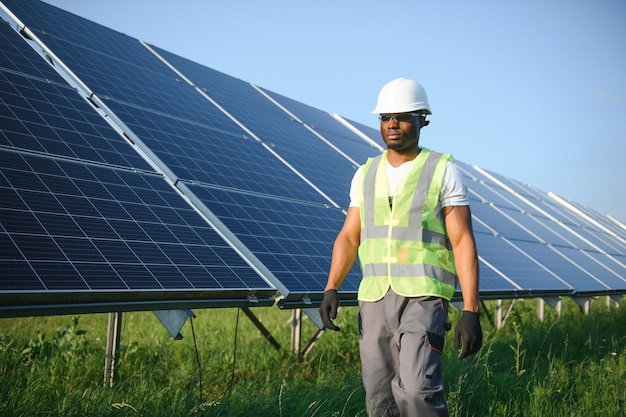 Homme afro-américain en casque blanc et combinaison grise debout parmi les rangées de panneaux solaires