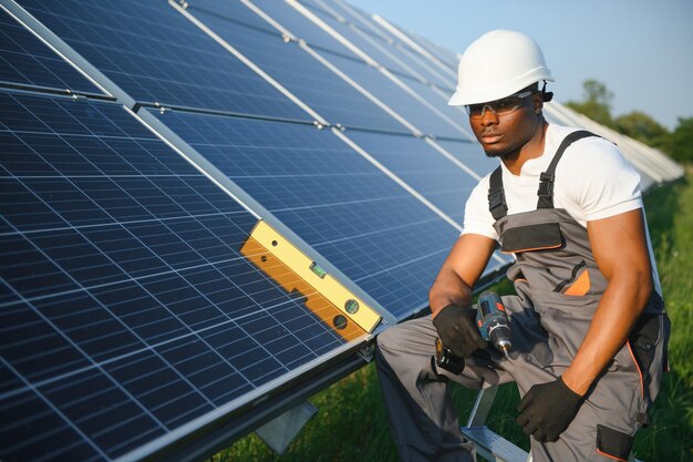 Homme afro-américain en casque blanc et salopette grise debout parmi des rangées de panneaux solaires