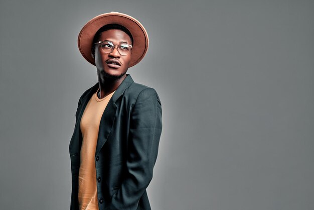 Homme afro-américain en chapeau à la mode de lunettes de soleil et veste décontractée regarde de côté fond gris isolé