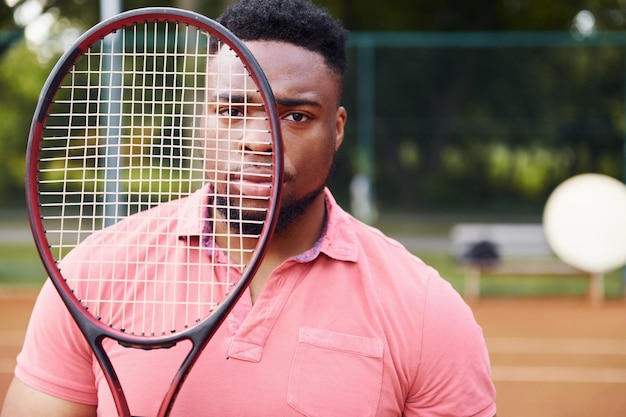 Photo homme afro-américain en chemise rose debout avec une raquette de tennis sur le terrain à l'extérieur