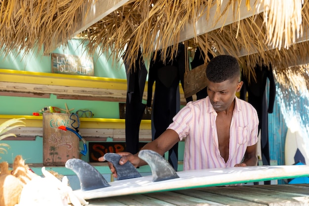 Homme afro-américain concentré préparant une planche de surf derrière le comptoir d'une cabane de plage de location de surf. Entreprise locale, propriétaire d'entreprise, loisirs, sport, surf, été et vacances, inchangés.