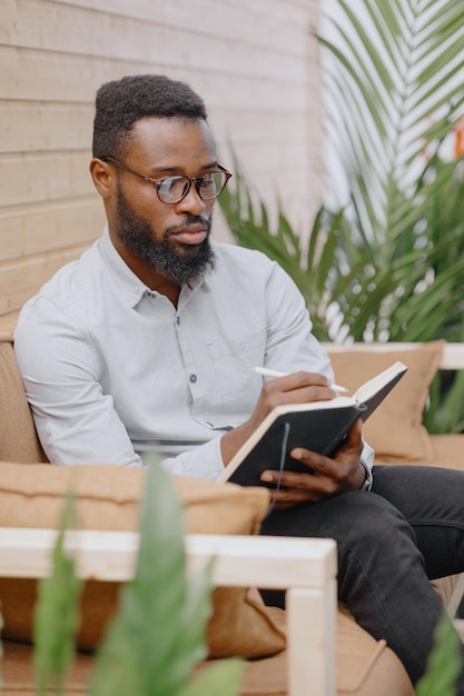 Un homme afro-américain dans un bureau ou un coworking prend des notes dans un cahier et travaille sur un ordinateur portable