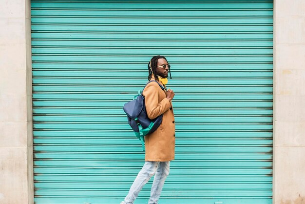 Homme afro-américain en dreadlocks avec lunettes de soleil élégantes et sac à dos marchant sur fond bleu