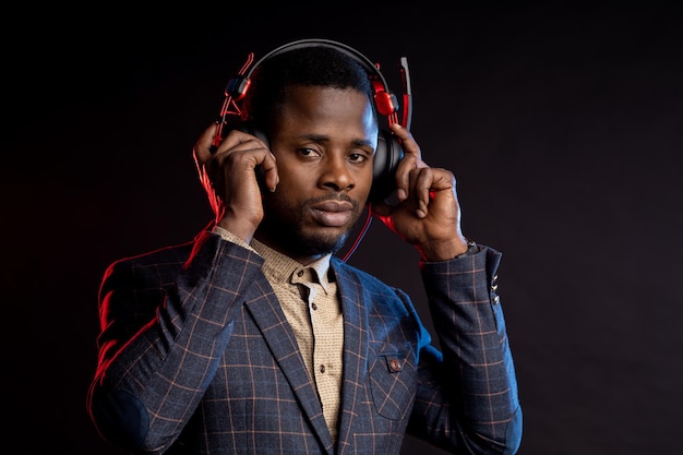 Homme afro-américain écoutant de la musique dans des écouteurs dans l'obscurité. Portrait de DJ élégant avec casque, vêtu d'un costume à carreaux, isolé sur fond noir