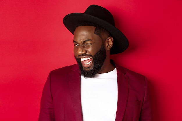 Homme afro-américain effronté en tenue de fête, faisant un clin d'œil à la caméra et souriant, debout sur fond rouge.