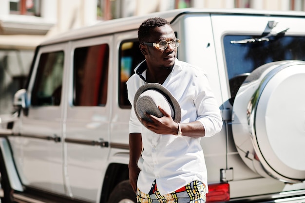 Homme afro-américain élégant en chemise blanche et pantalon coloré avec chapeau et lunettes posés en plein air contre la voiture suv de la mafia argentée Garçon modèle à la mode noir