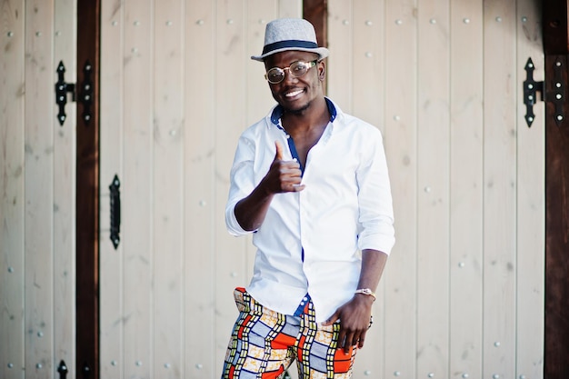 Homme afro-américain élégant en chemise blanche et pantalon coloré avec chapeau et lunettes posés en plein air garçon modèle à la mode noir