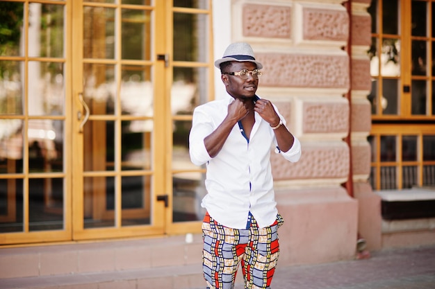 Homme afro-américain élégant en chemise blanche et pantalon coloré avec chapeau et lunettes posés en plein air garçon modèle à la mode noir