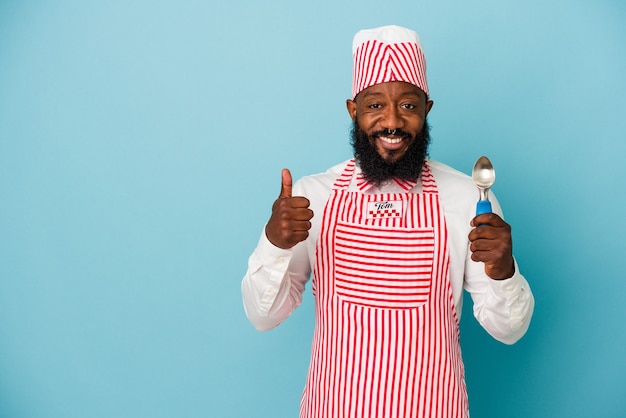 Homme afro-américain de fabricant de crème glacée tenant une boule de crème glacée d'isolement sur le fond bleu souriant et levant le pouce vers le haut