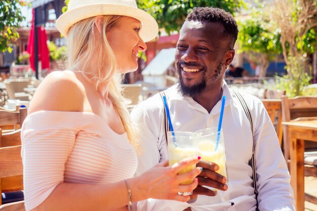 un homme afro-américain et une femme se regardent avec amour dans un café tropical d'été et boivent un cocktail de fruits frais à l'orange.