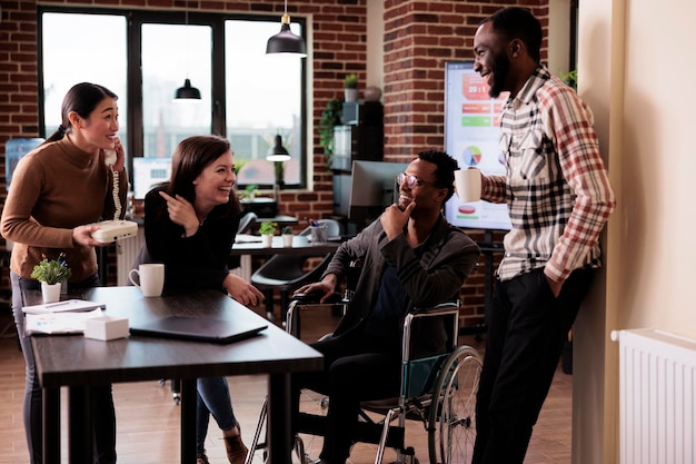 Homme afro-américain handicapé chronique riant avec ses collègues, travaillant dans un bureau adapté aux personnes handicapées. Des hommes d'affaires et des utilisateurs de fauteuils roulants discutent pendant une pause sur le lieu de travail de démarrage.