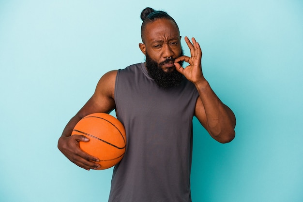 Homme afro-américain jouant au basket isolé sur un mur bleu avec les doigts sur les lèvres gardant un secret.