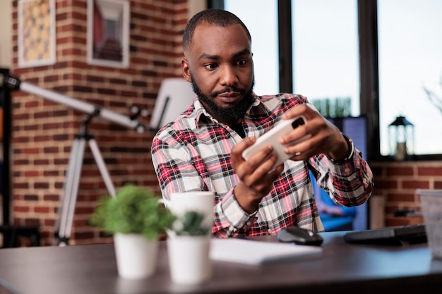 Homme afro-américain jouant à des jeux vidéo sur un téléphone portable, s'amusant avec une compétition de jeux en ligne et travaillant à domicile. Profitant du jeu sur smartphone, freelaner faisant des activités de loisirs.