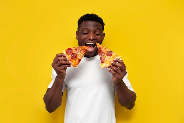 Un homme afro-américain joyeux en chemise blanche mange deux tranches de pizza avec la bouche ouverte.