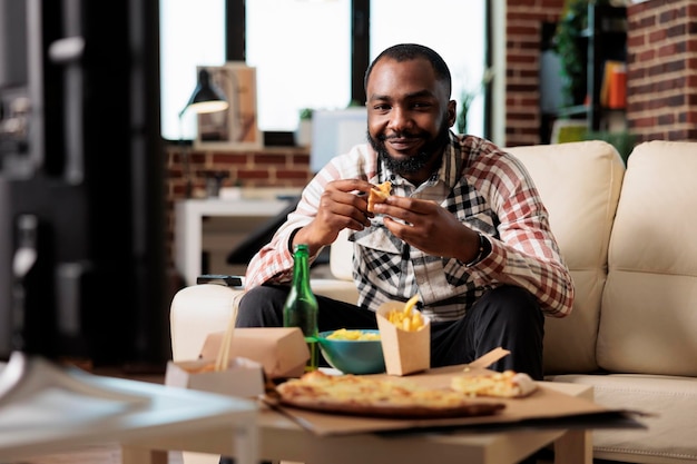 Homme afro-américain mangeant une tranche de pizza de la livraison à emporter pour regarder un film télévisé dans le salon. Regarder un film à la télévision avec un repas rapide à emporter, s'amuser avec des collations et de la bière.