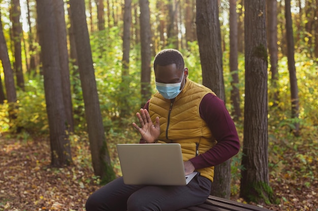 Homme afro-américain en masque médical assis dans le parc de la ville sur un banc avec appel vidéo pour ordinateur portable avec ven