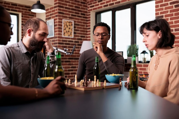 Homme afro-américain pensif pensant au prochain mouvement d'échecs assis à table. Personne concentrée et confiante assise à la maison dans le salon avec des amis multiethniques tout en jouant à des jeux de stratégie.