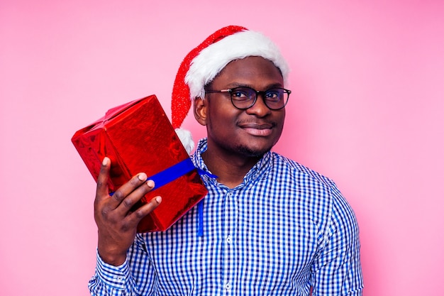 Homme afro-américain portant une chemise à carreaux élégante grand sourire en bonnet de Noel avec boîte-cadeau sur fond rose studio
