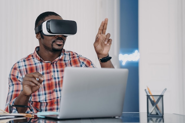 Homme afro-américain portant des lunettes VR travaillant sur un ordinateur portable en réalité virtuelle assis au bureau