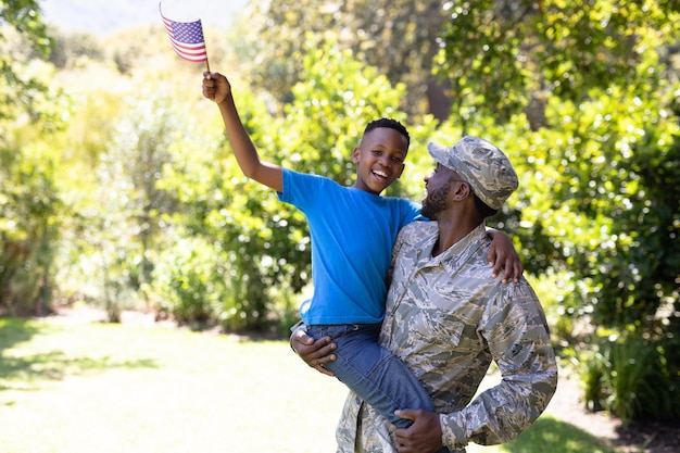 Homme afro-américain portant un uniforme militaire, rentrant chez lui, tenant son fils, embrassant, regardant la caméra et souriant, un garçon tient un mini drapeau, par une journée ensoleillée