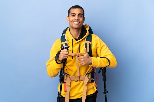 Homme afro-américain avec sac à dos et bâtons de randonnée sur un mur isolé donnant un coup de pouce vers le haut