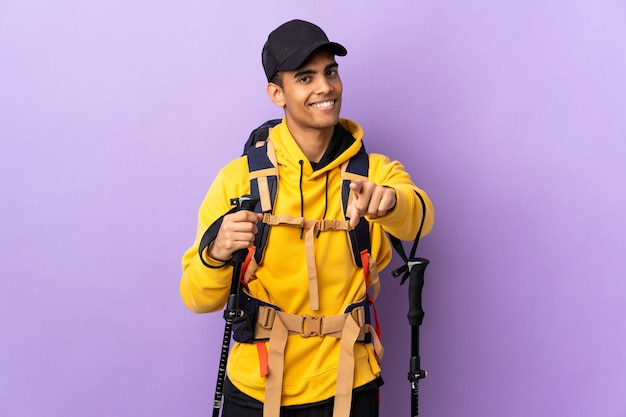 Homme afro-américain avec sac à dos et bâtons de randonnée sur le mur isolé pointant vers l'avant avec une expression heureuse