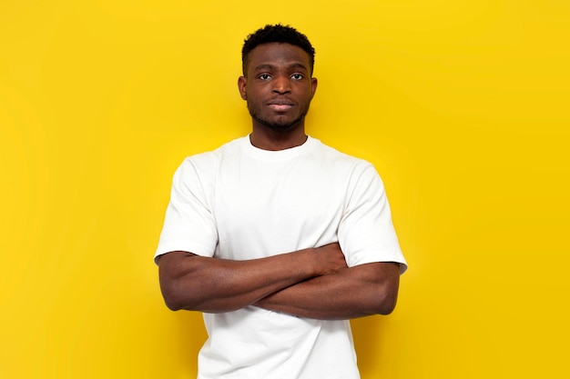 Un homme afro-américain sérieux en T-shirt blanc se tient avec les bras croisés sur un fond jaune