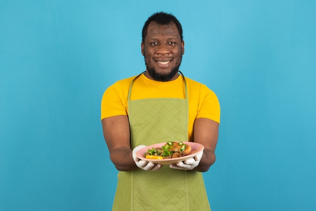 Un homme afro-américain souriant à la barbe, portant un tablier avec une assiette de nourriture dans les mains, se dresse sur le mur bleu.