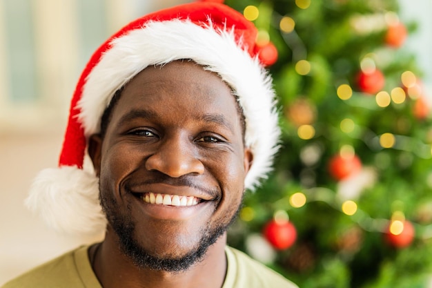 Homme afro-américain avec un sourire blanc comme neige en bonnet de noel regardant la caméra près de l'arbre de noël à la maison