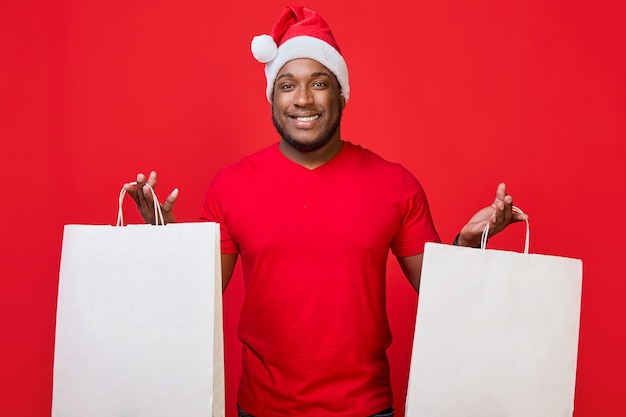 L'homme afro-américain sourit dans un bonnet de Noel et tient des sacs en papier blanc sur fond rouge