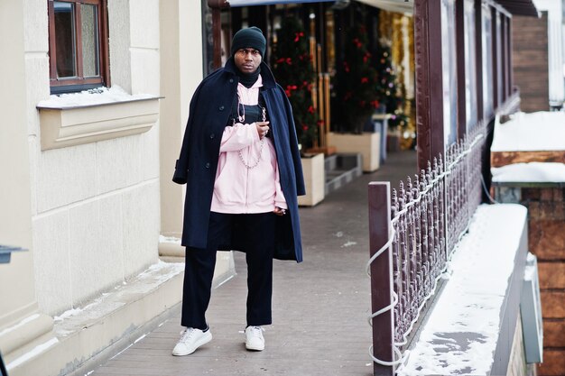 Homme afro-américain de style urbain élégant en manteau noir et chapeau à capuche rose posé le jour de l'hiver