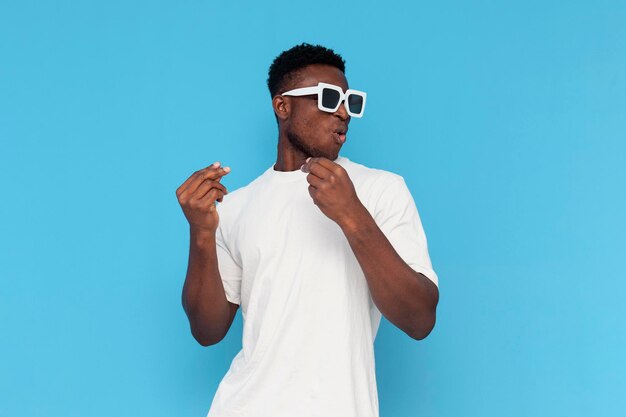 homme afro-américain en t-shirt blanc et lunettes de soleil danse sur de la musique et chante sur fond bleu