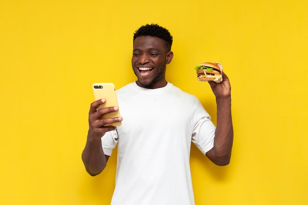 Un homme afro-américain en T-shirt blanc tient un grand hamburger et utilise un smartphone sur un fond jaune