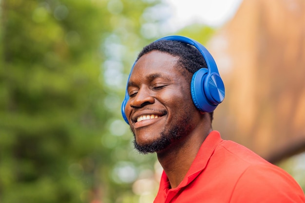 Homme afro-américain en t-shirt rouge écoutant de la musique dans des écouteurs bleus et étant de bonne humeur à l'extérieur