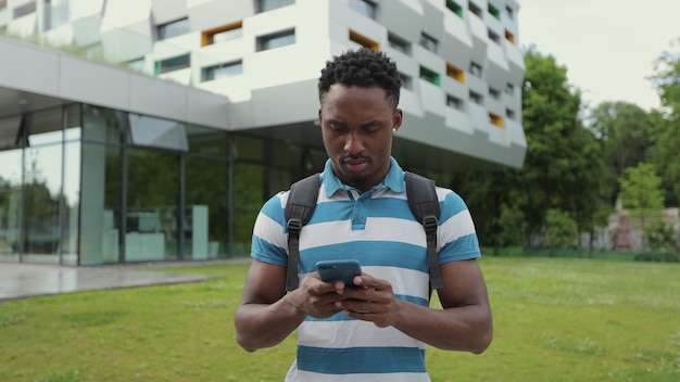 Homme afro-américain utilisant un smartphone sur le fond de l'homme universitaire lisant et tapant un message au téléphone à l'aide d'une application souriant heureux qu'il se tenait près du centre de bureau dans la rue