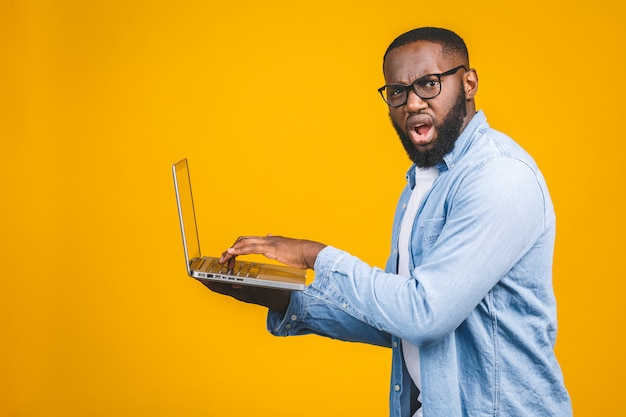 Homme afro-américain ypung effrayé une mauvaise nouvelle sur son ordinateur portable, isolé sur fond jaune.