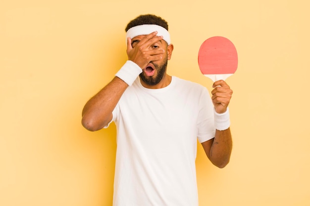 Homme afro noir regardant choqué effrayé ou terrifié couvrant le visage avec le concept de ping-pong à la main