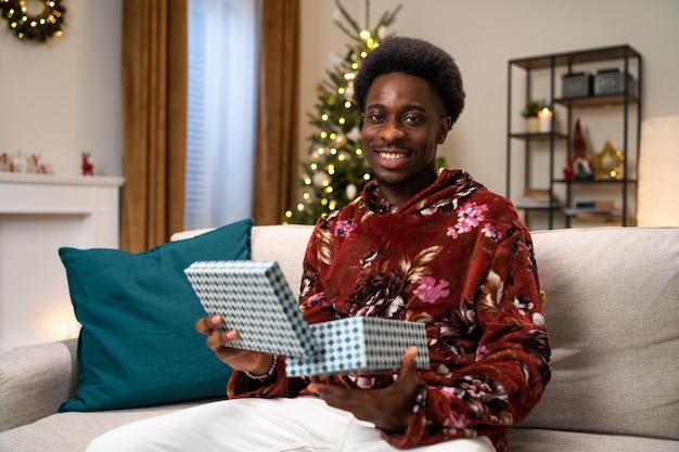 Un homme afro souriant est assis sur un canapé à côté d'un arbre de noël, il ouvre un cadeau