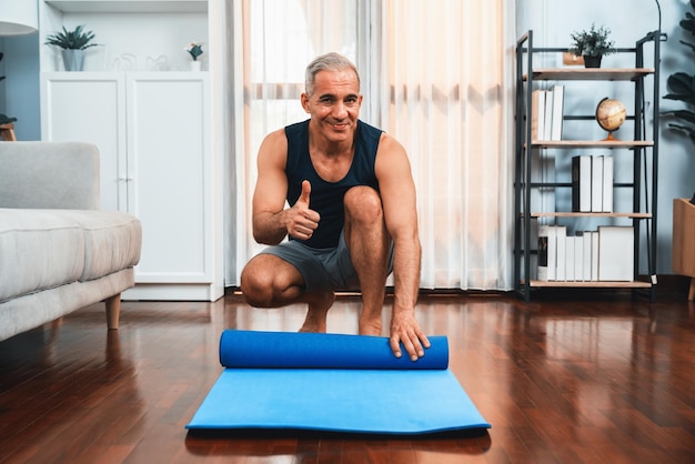 Homme âgé actif et sportif préparant un tapis d'exercice de remise en forme roulant sur le sol du salon à la maison Exercice à domicile comme concept de mode de vie corporel sain après la retraite Clout