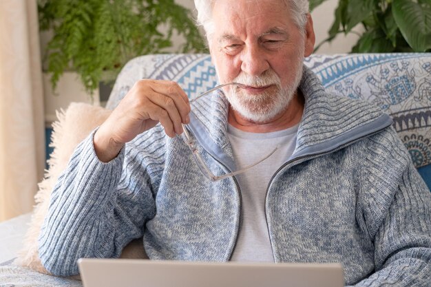 Homme âgé des années 70 assis sur un canapé naviguant sur un ordinateur portable senior regardant un écran d'ordinateur portable surfant sur le net ancienne génération et application technologique moderne concept d'utilisation facile