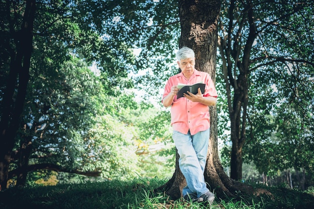 Homme âgé asiatique debout sous un arbre dans le parc et lit un livre de journal