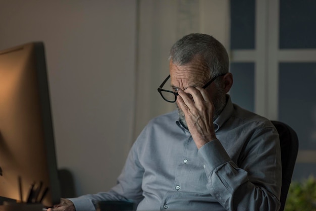 Un homme âgé assis à son bureau et travaillant avec son ordinateur tard dans la nuit il ajuste ses lunettes